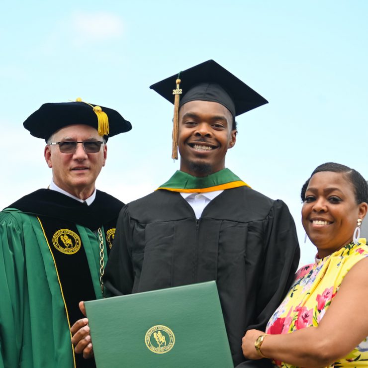 Methodist University student poses with a family member and President Stanley T. Wearden at a spring commencement graduation at Segra Stadium