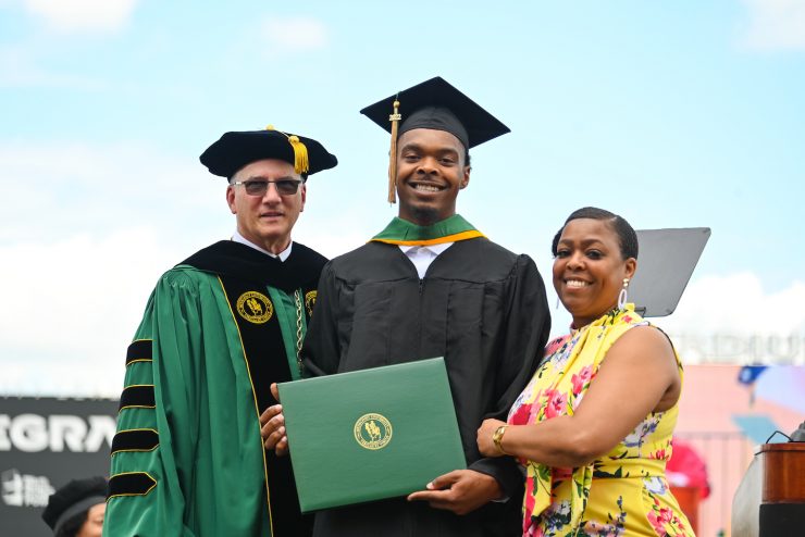 Methodist University student poses with a family member and President Stanley T. Wearden at a spring commencement graduation at Segra Stadium