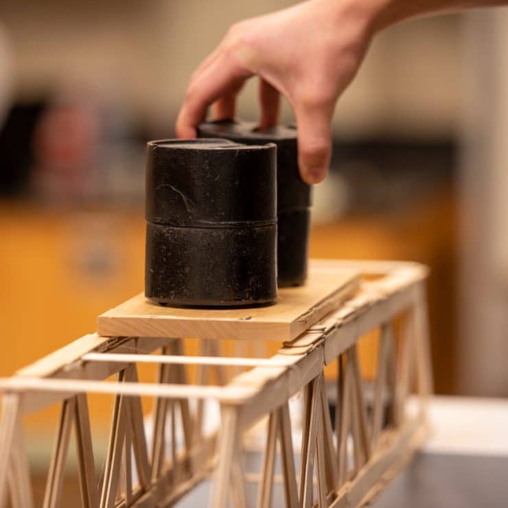 A student places a weight on their bridge to test the sturdiness of their project