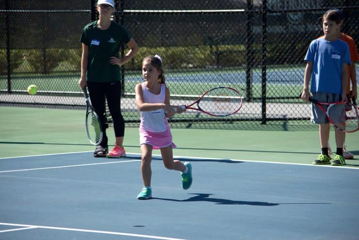 Counselors and participants watch as a youth camp participant prepares to swing at a ball with her racquet.